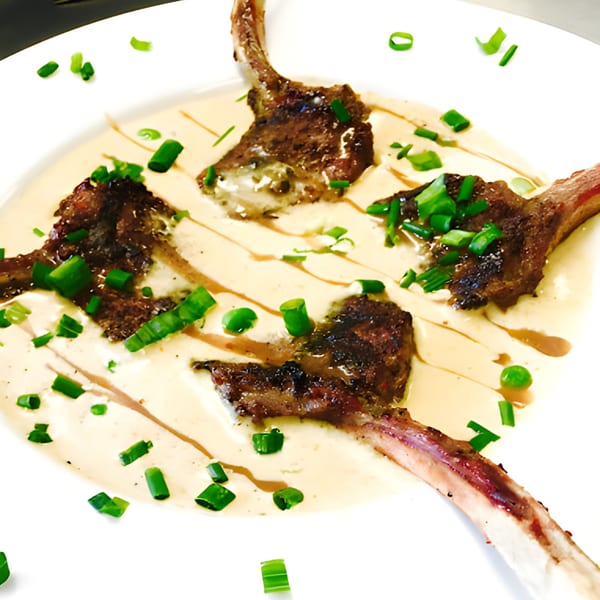 Four lamb chops on a white plate, drizzled with a creamy sauce and garnished with green onions.