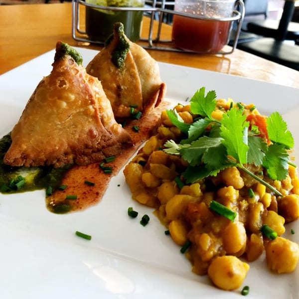 Two golden-brown samosas with green chutney, alongside chickpea curry and fresh cilantro on a white plate.